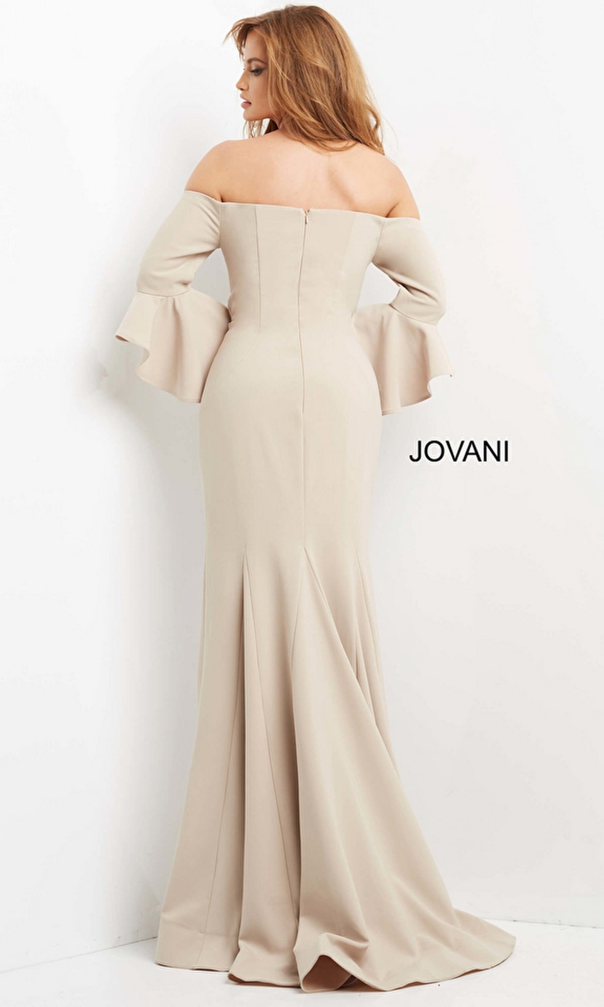 A woman wearing the Jovani 07065 Bell-Sleeve Designer Evening Gown stands facing a white wall. The floor-length beige dress showcases timeless elegance, with
